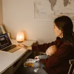 A mother working on a laptop while holding her sleeping baby, creating a warm at-home atmosphere.