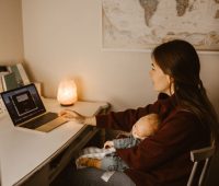 A mother working on a laptop while holding her sleeping baby, creating a warm at-home atmosphere.