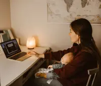 A mother working on a laptop while holding her sleeping baby, creating a warm at-home atmosphere.